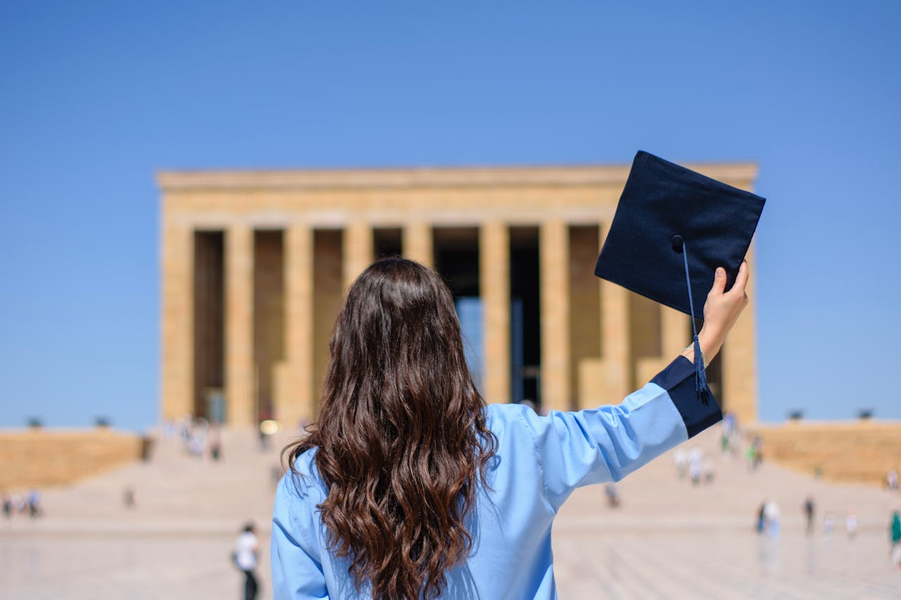 A woman celebrates graduation at Anıtkabir, holding a cap against a clear summer sky.