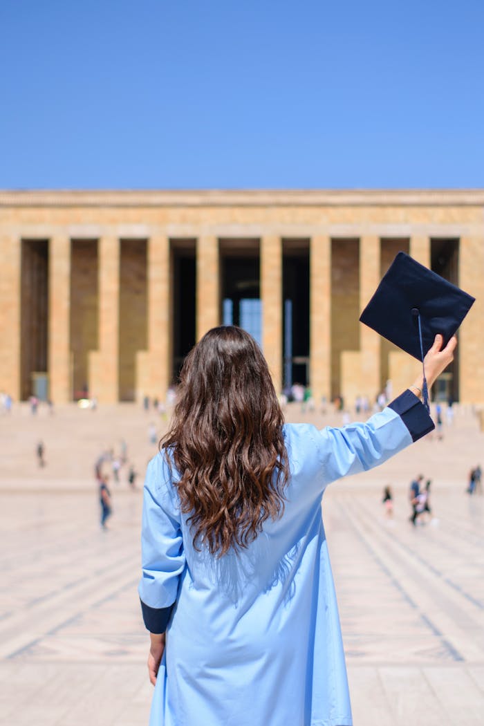 Young graduate in blue cap and gown celebrating outside a historic monument on a sunny day.
